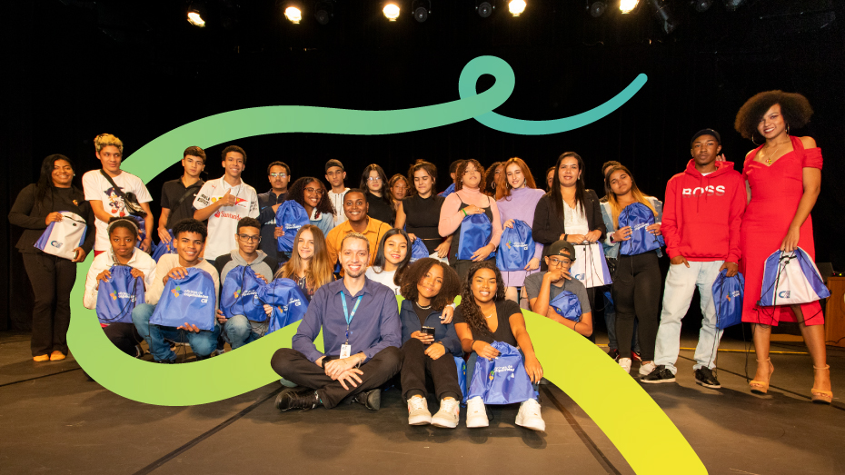 Foto de um grupo de jovens em cima de um palco sorrindo. Cada um segurando uma mochila azul.